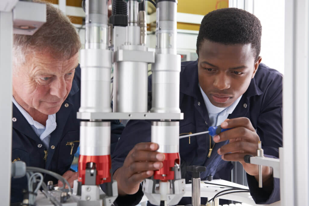 Engineer And Apprentice Working On Machine In Factory